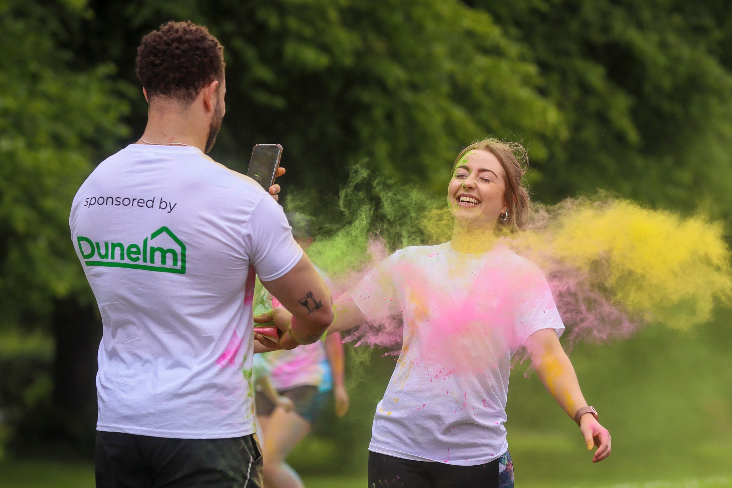 Man wearing a tshirt that says sponsored by Dunelm takes a photograph of a smiling girl being covered in paint powder at Inspire's colour run