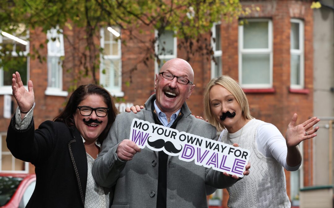 Three people standing outside a building, two women wearing fake moustaces beside a man holding a sign saying mo your own way for woodvale
